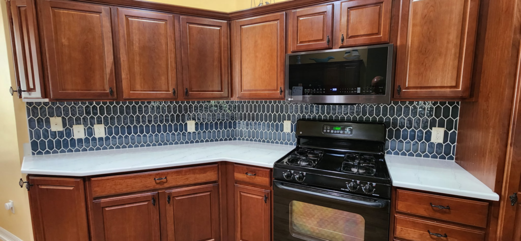 Custom backsplash installation featuring unique dark geometric hexagon tiles in a warm kitchen with wood cabinets and a gas range.