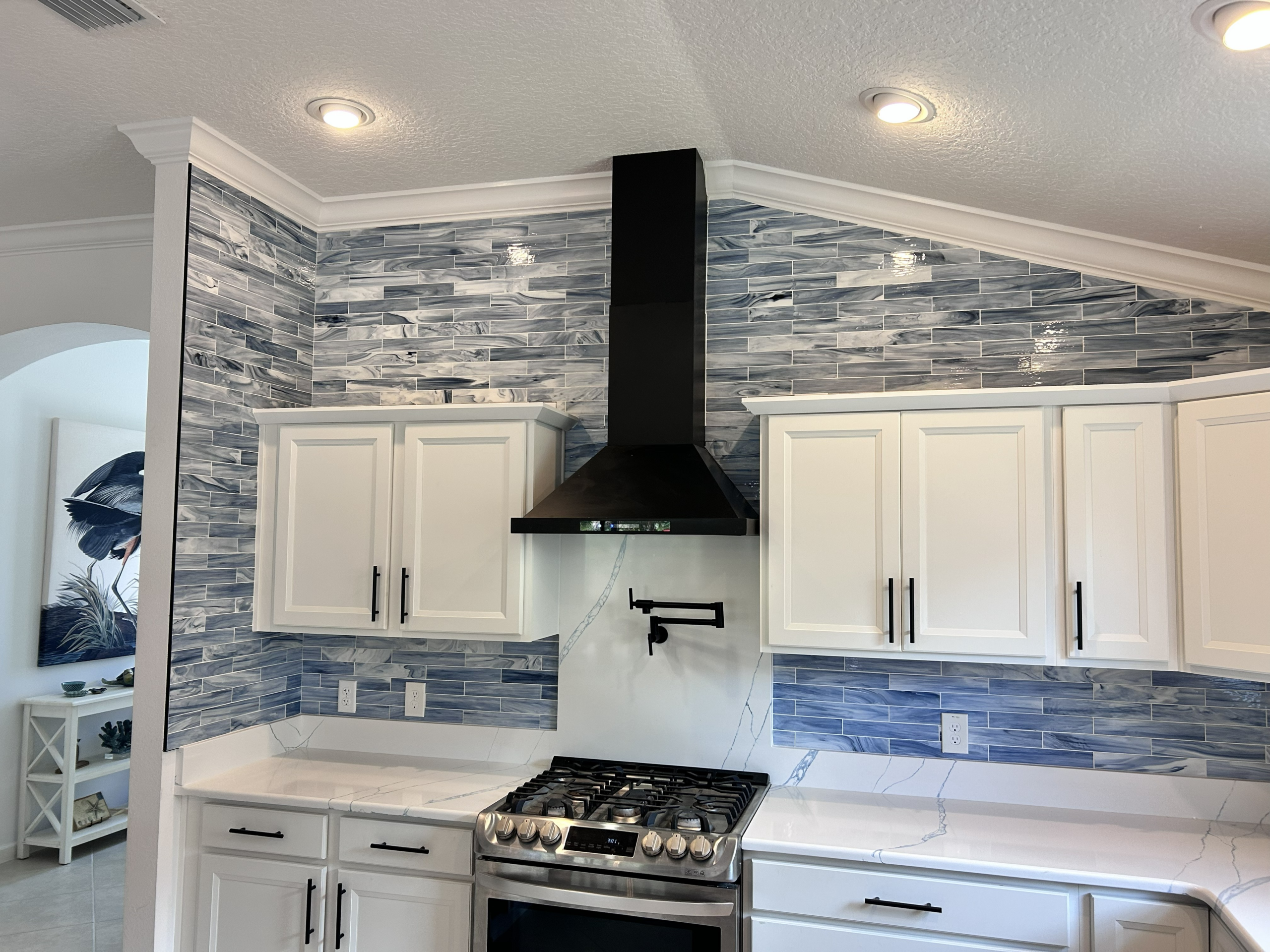Full-wall blue glass subway tile backsplash installation featuring a black vent hood and pot filler by CRB Services Pro.